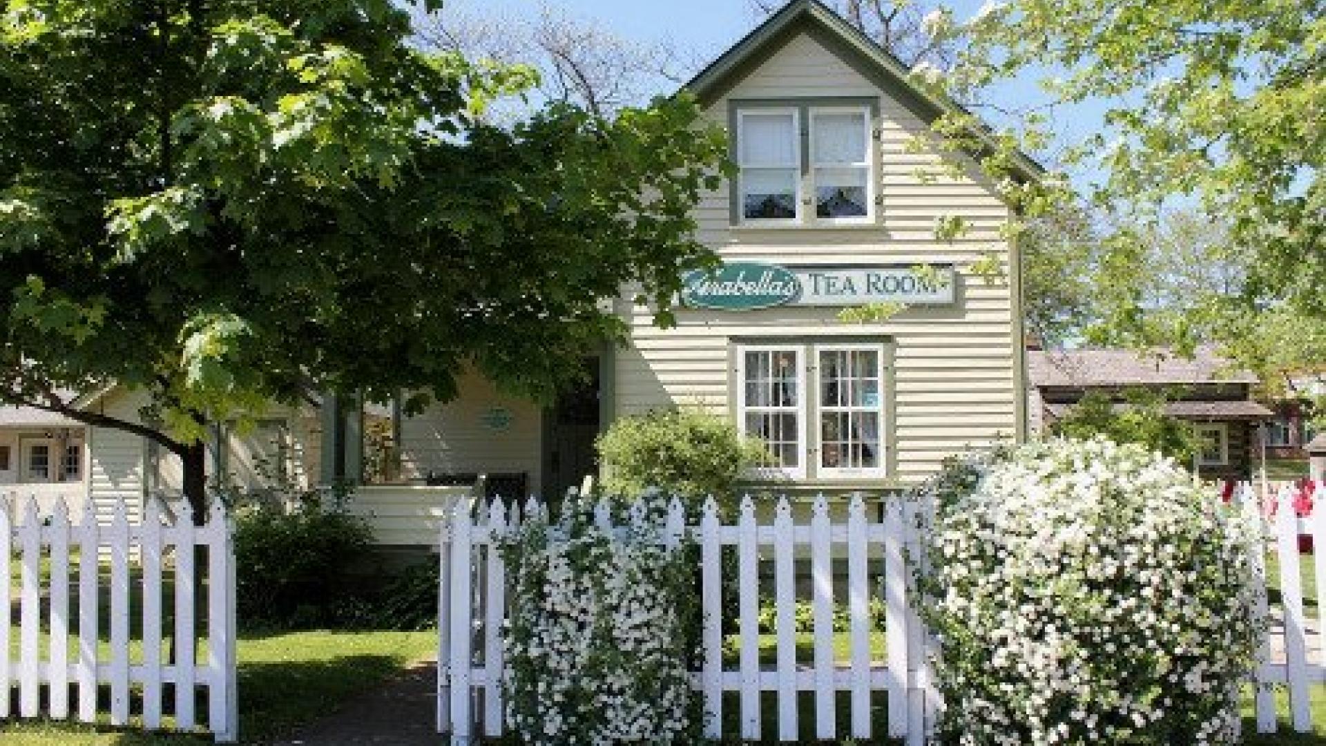 Cozy house behind a white picket fence with lush greenery and blooming bushes.