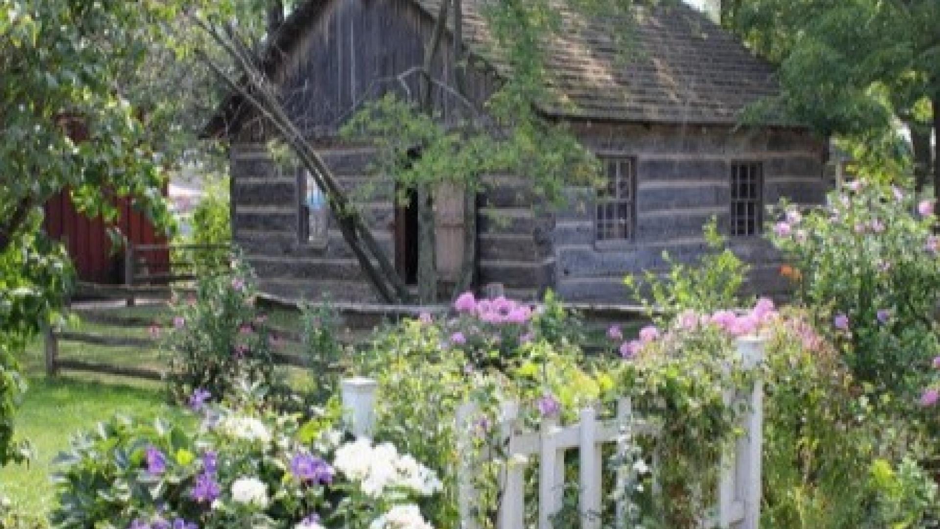 Log cabin behind a blooming flower garden and white picket fence.