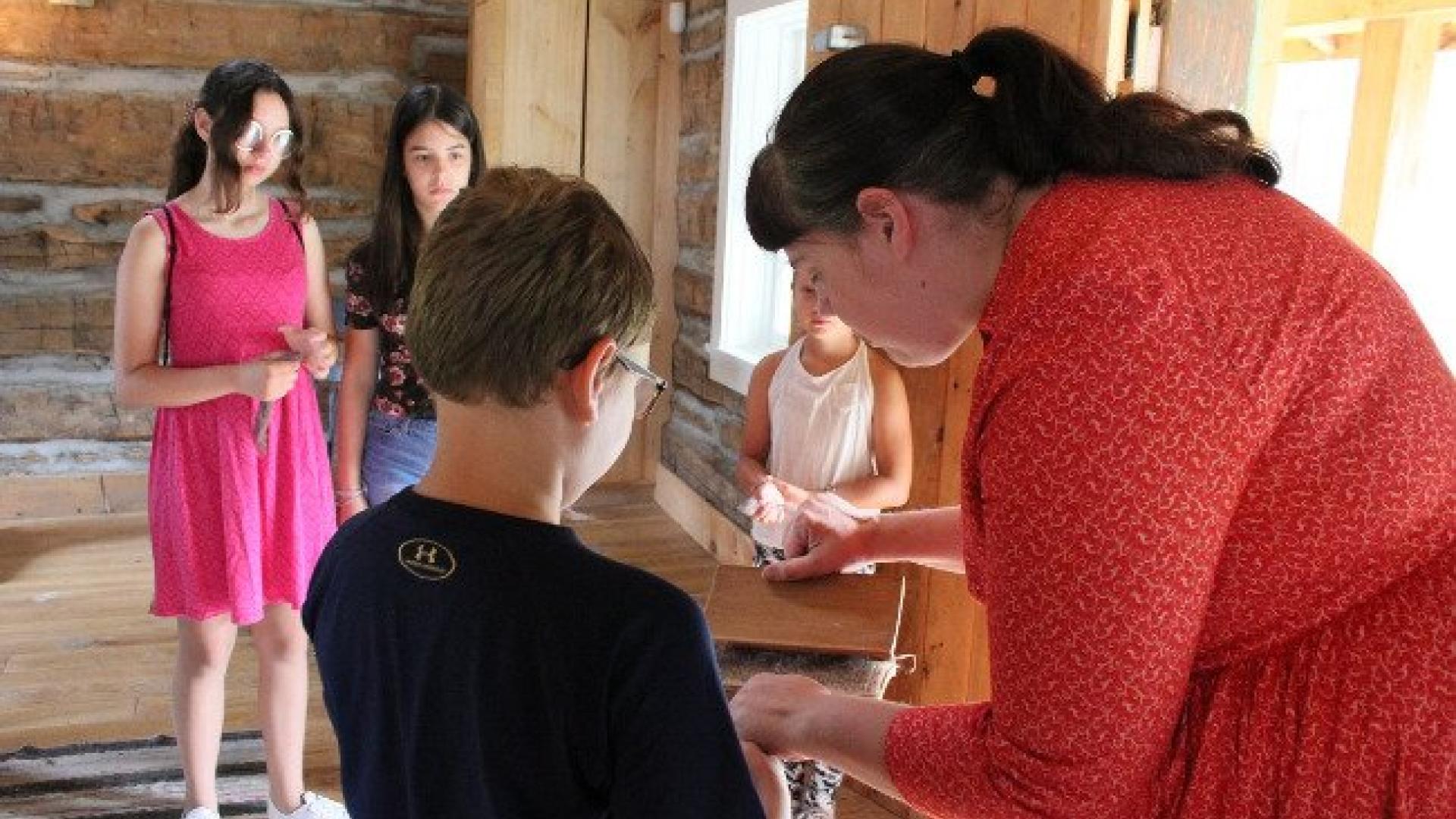 Woman in red shows craft to children in a wooden cabin.