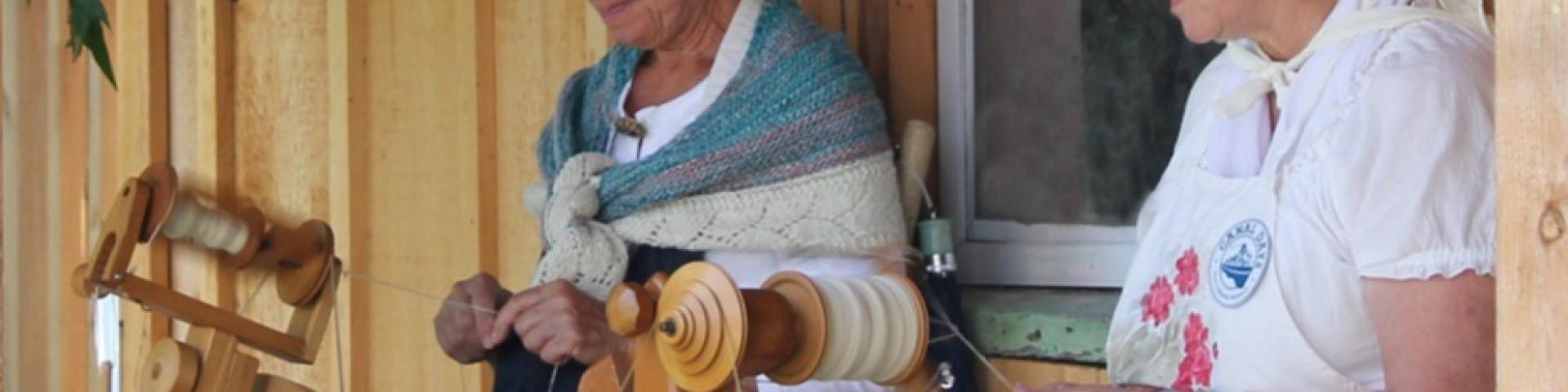 Two people spinning yarn on wooden spinning wheels on a porch.