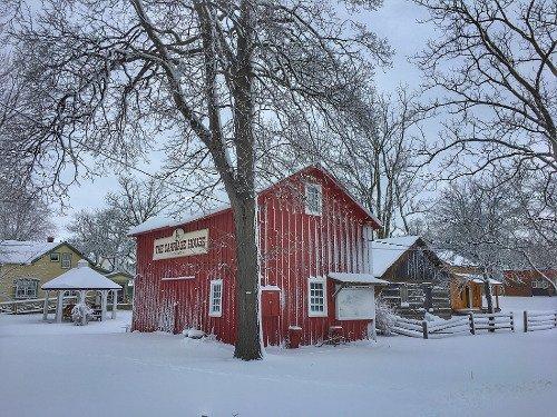 Red barn in snowy landscape with bare trees and houses nearby.