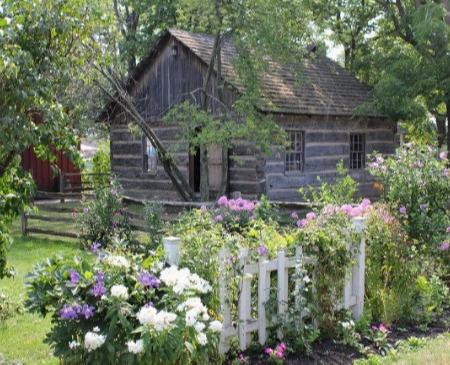 Log cabin behind a blooming flower garden and white picket fence.