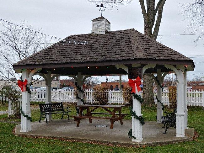 Gazebo decorated with wreaths and bows, empty benches, overcast sky.
