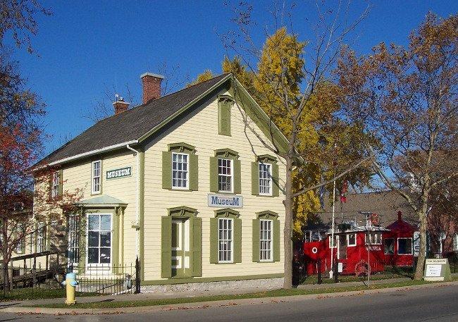 Yellow historic building with green shutters, blue sky background. Large tree nearby.