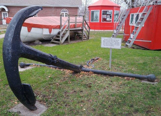 Large black anchor displayed on grass near a red building.