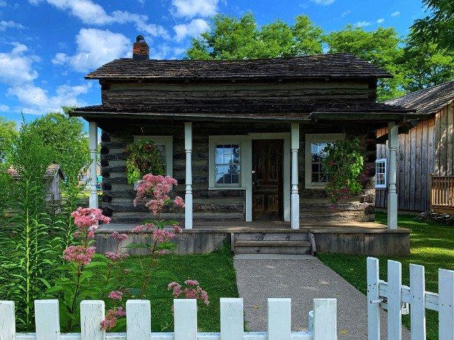 Rustic log cabin with a garden and white picket fence.