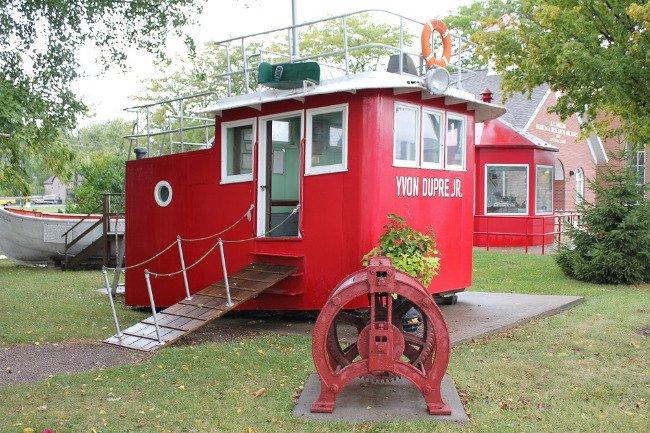 Red tugboat replica with a wooden ramp on grass, surrounded by trees and buildings.