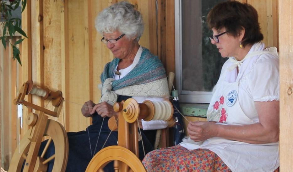 Two people spinning yarn on wooden spinning wheels on a porch.