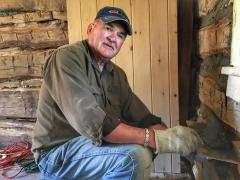 Man plastering a log cabin wall, wearing gloves and a cap.