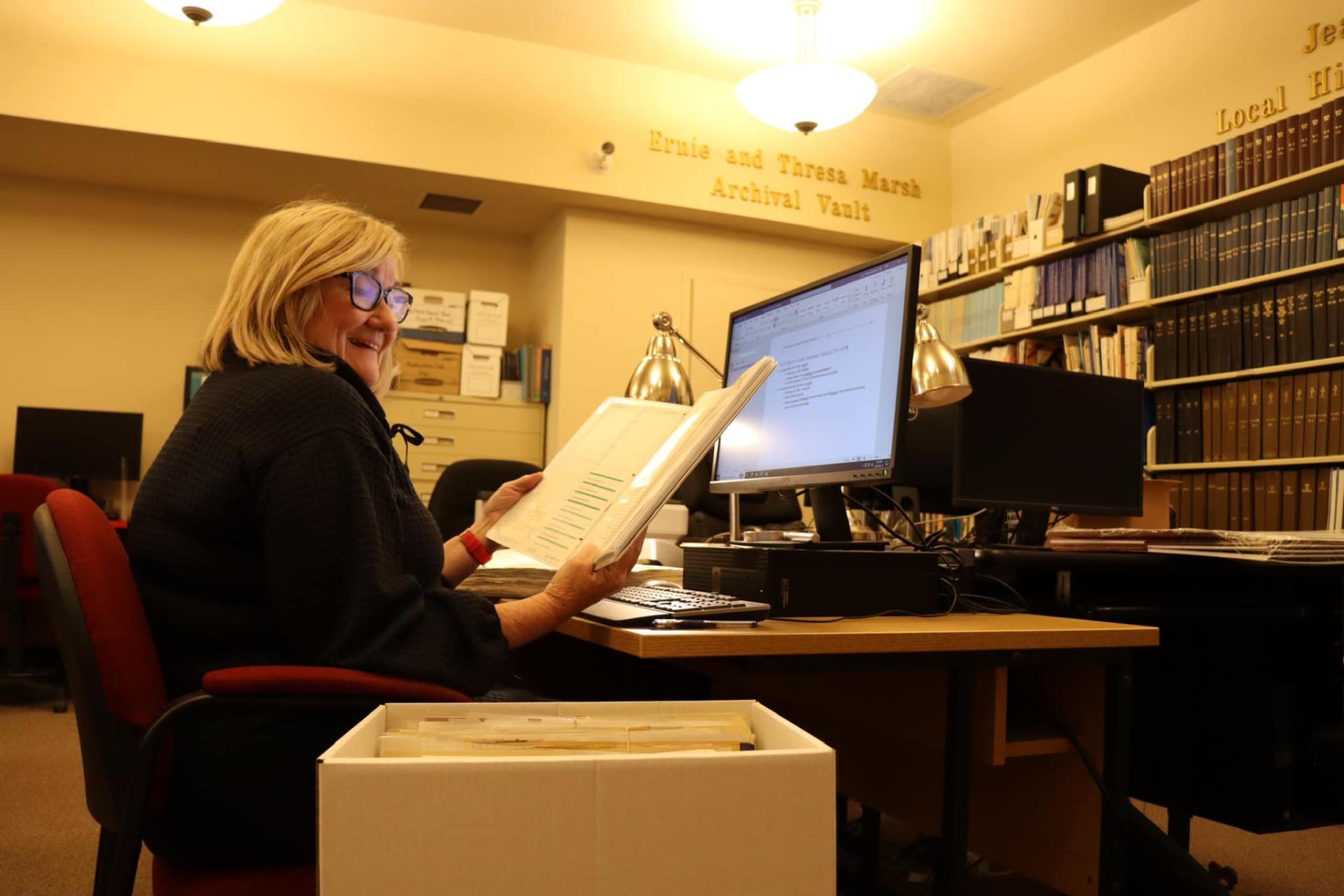 Person reading a document at a desk in a library with books and a computer.